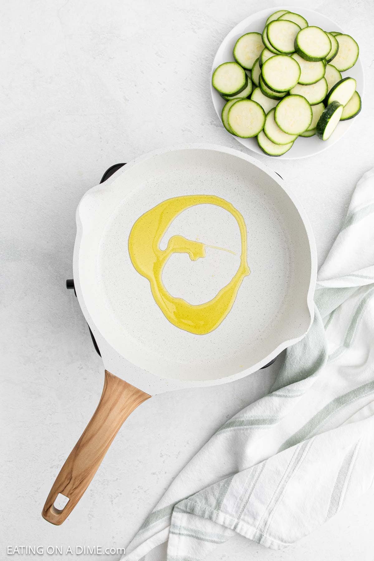 A white skillet with oil rests on a light surface. Its wooden handle extends gracefully, while beside it, a small white plate holds pan-fried zucchini slices. A green and white striped cloth complements the scene perfectly.