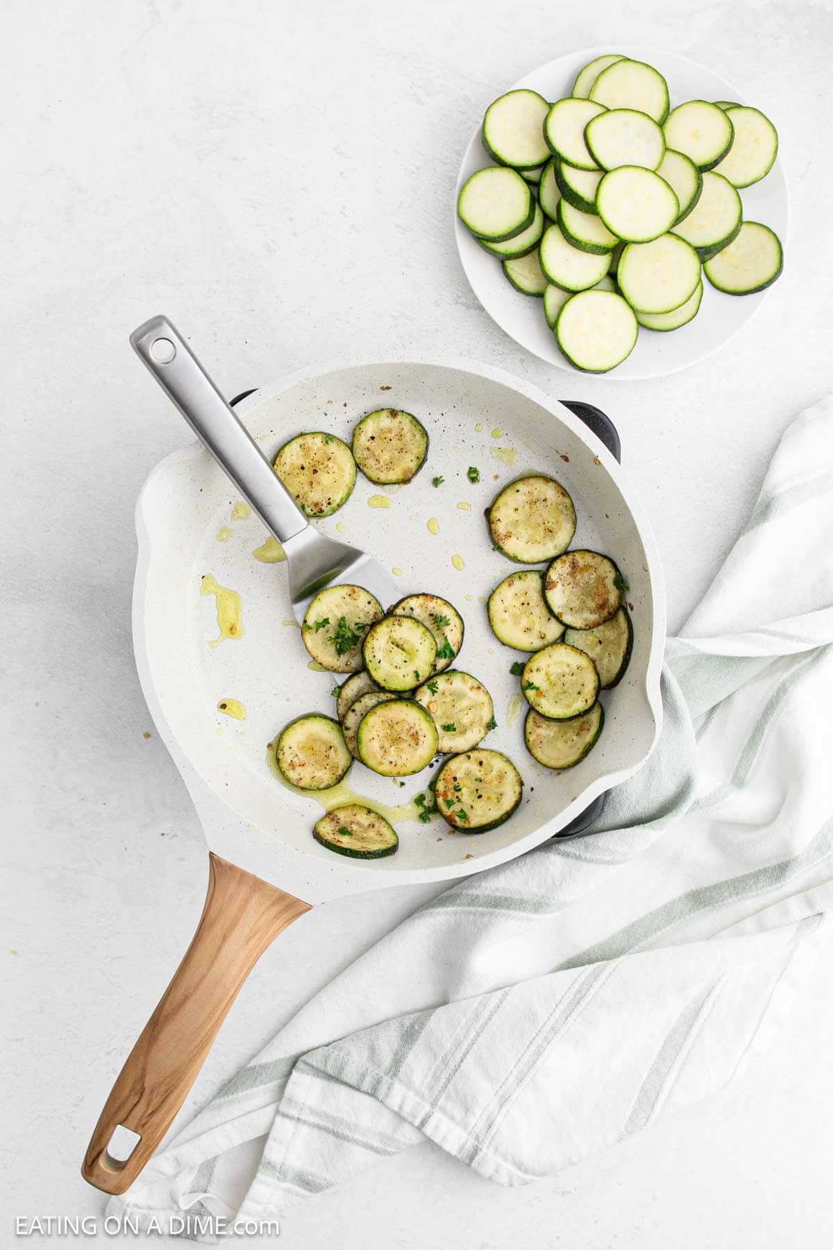 A skillet with pan-fried zucchini slices on a white surface, accompanied by a wooden-handled spatula. A white plate with raw zucchini slices sits beside the skillet. A folded striped towel is also partially visible.