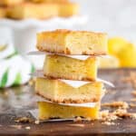 A stack of four lemon bars from an easy lemon bars recipe sits on a wooden surface, separated by parchment paper, with crumbs scattered around and a blurred lemon and dessert tray in the background.