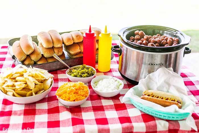 A hot dog bar set up on an outside table.  Chips, hot dog buns, cheese, relish, onions, ketchup, mustard and a small crock pot full of hot dogs are on the table. 
