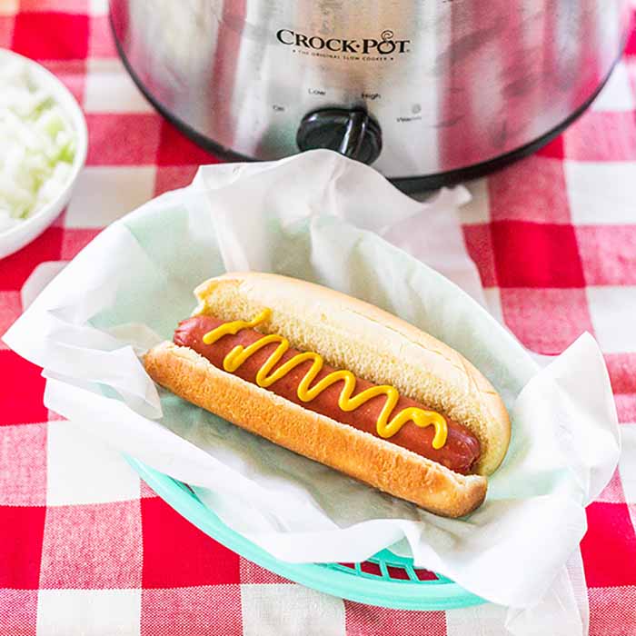 A hot dog with mustard on it in front of a small crock pot full of hot dogs 