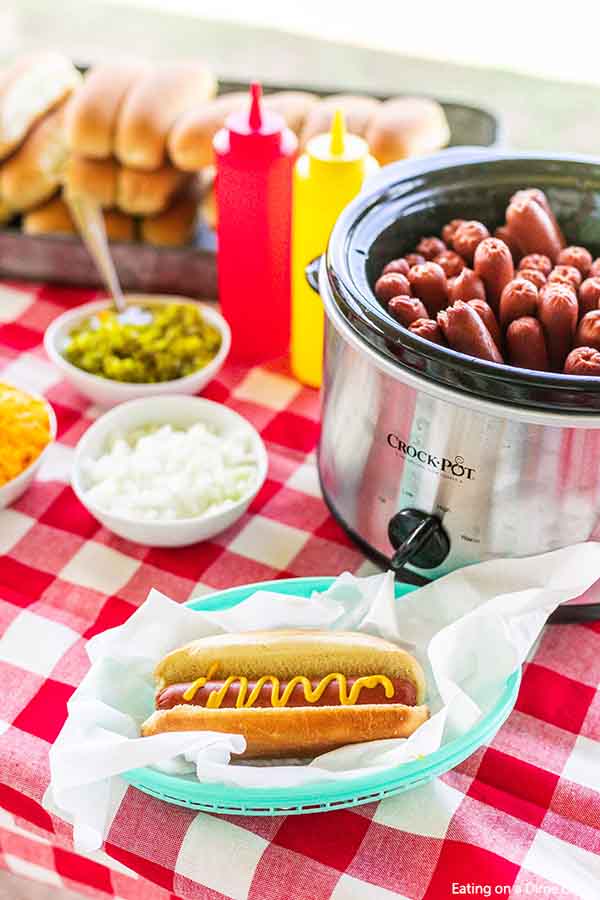 A hot dog with mustard on it in front of a small crock pot full of hot dogs 