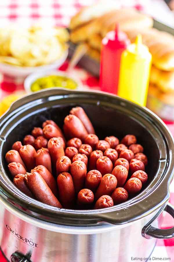 Hot Dogs being cooked in a crock pot with other hot dog condiments behind it. 
