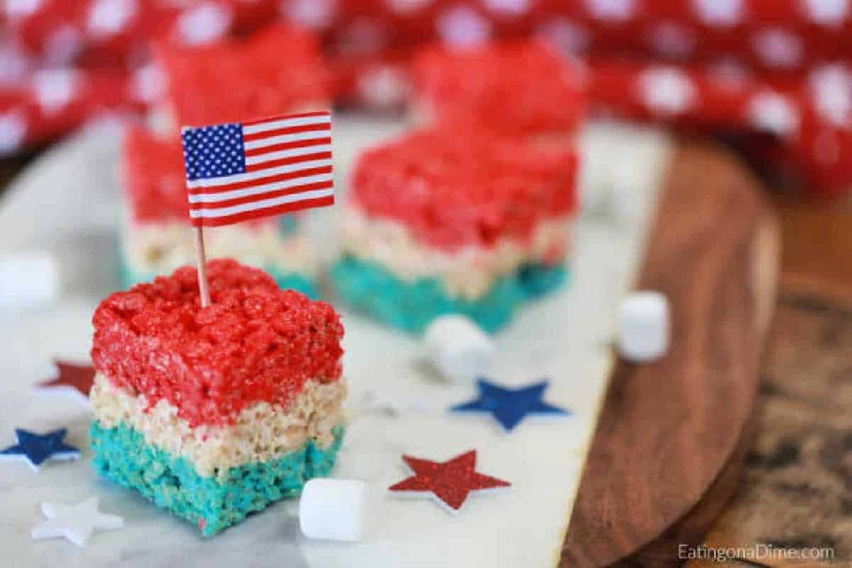 Red, white, and blue Rice Krispie Treats and topped with a small American flag, displayed on a marble surface with star decorations.