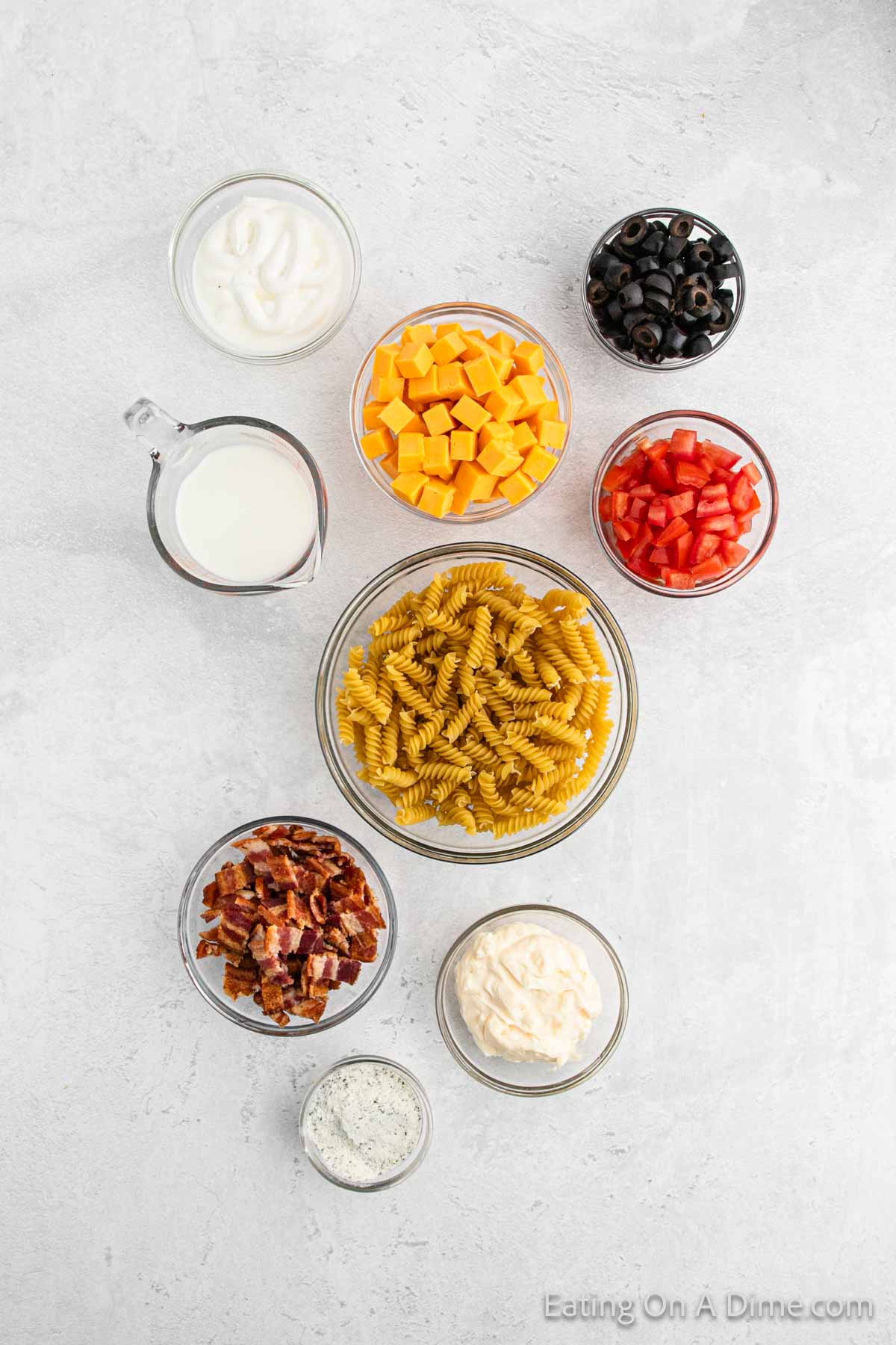 Bowls of pasta, ranch, black olives, cheese cubes, diced tomatoes, chopped bacon, mayonnaise, sour cream