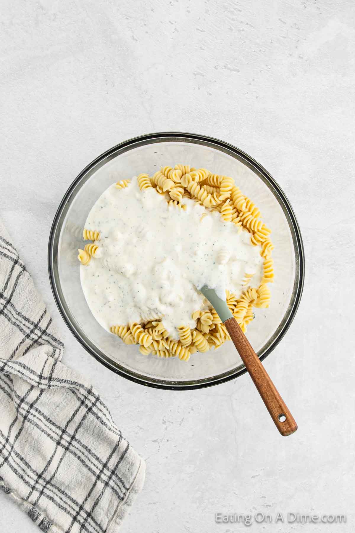 Pouring the ranch dressing over the pasta salad in a bowl with a spoon