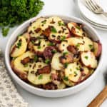 german potato salad served in a white bowl, parsley, plates and forks blurred in the background.