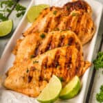 Grilled chicken breasts arranged on a white rectangular platter, marinated in a zesty chicken taco marinade, garnished with chopped parsley and served with lime wedges. Fresh parsley and lime slices are also placed around the plate.