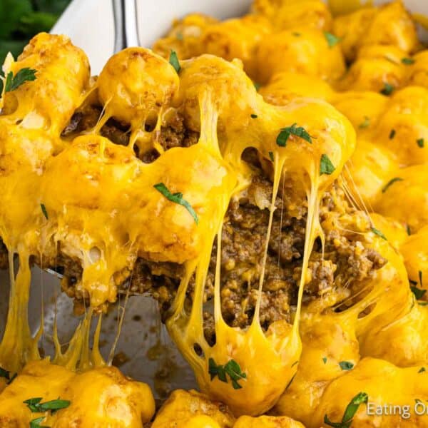 A serving of cheeseburger tator tot casserole is being scooped from a baking dish, revealing melted cheddar cheese, ground beef, and crispy tater tots, all garnished with chopped parsley.
