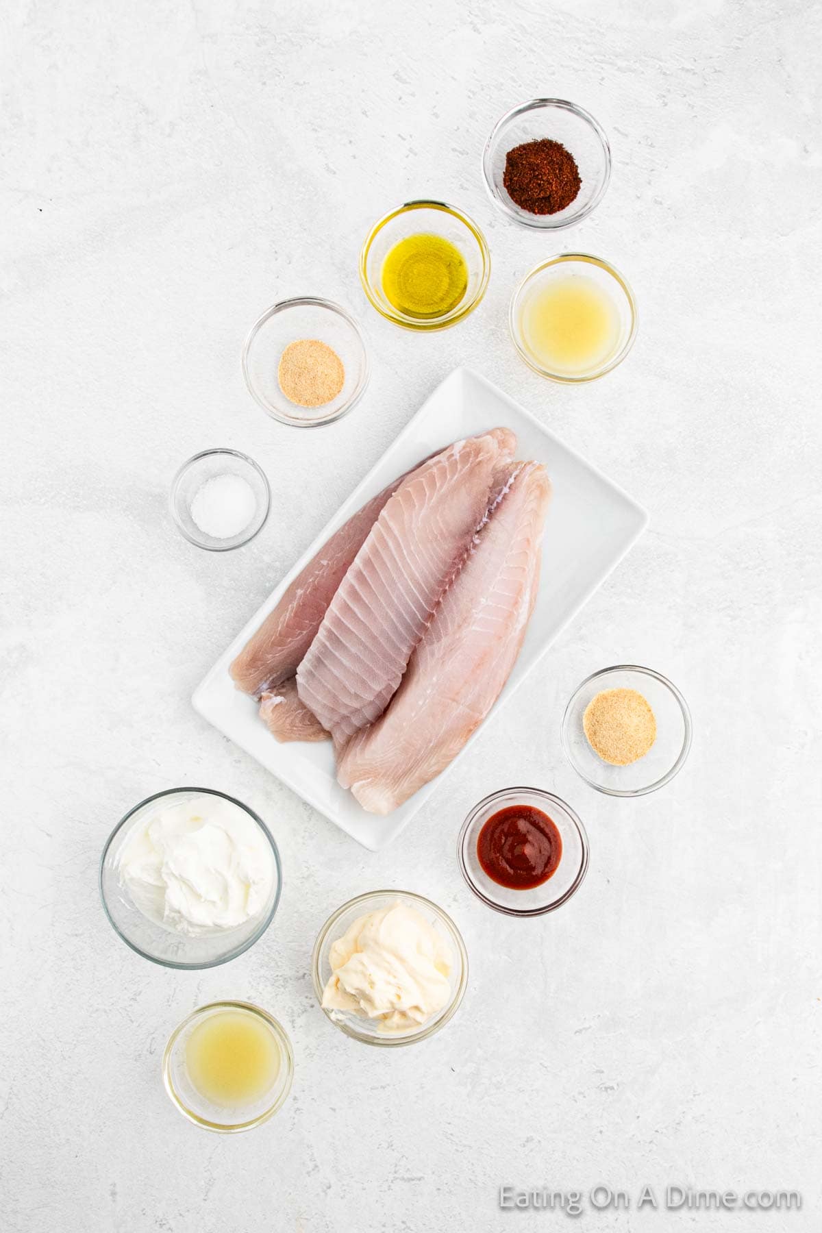 Top view of two uncooked fish fillets on a rectangular white plate surrounded by various small bowls containing olive oil, seasonings, yogurt, mayonnaise, spice powder, lemon juice, and a small amount of pink sauce—everything you need for an easy fish tacos recipe—on a light gray surface.