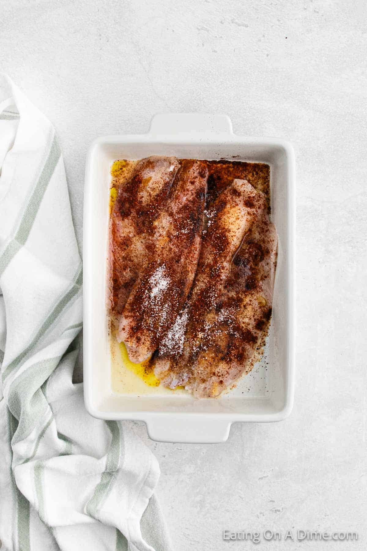 A white rectangular baking dish contains raw, seasoned chicken breasts. The chicken is sprinkled with spices, including paprika, black pepper, and salt. Next to the baking dish is a white and green striped kitchen towel on a light gray surface—a perfect scene reminiscent of preparing an easy fish tacos recipe.