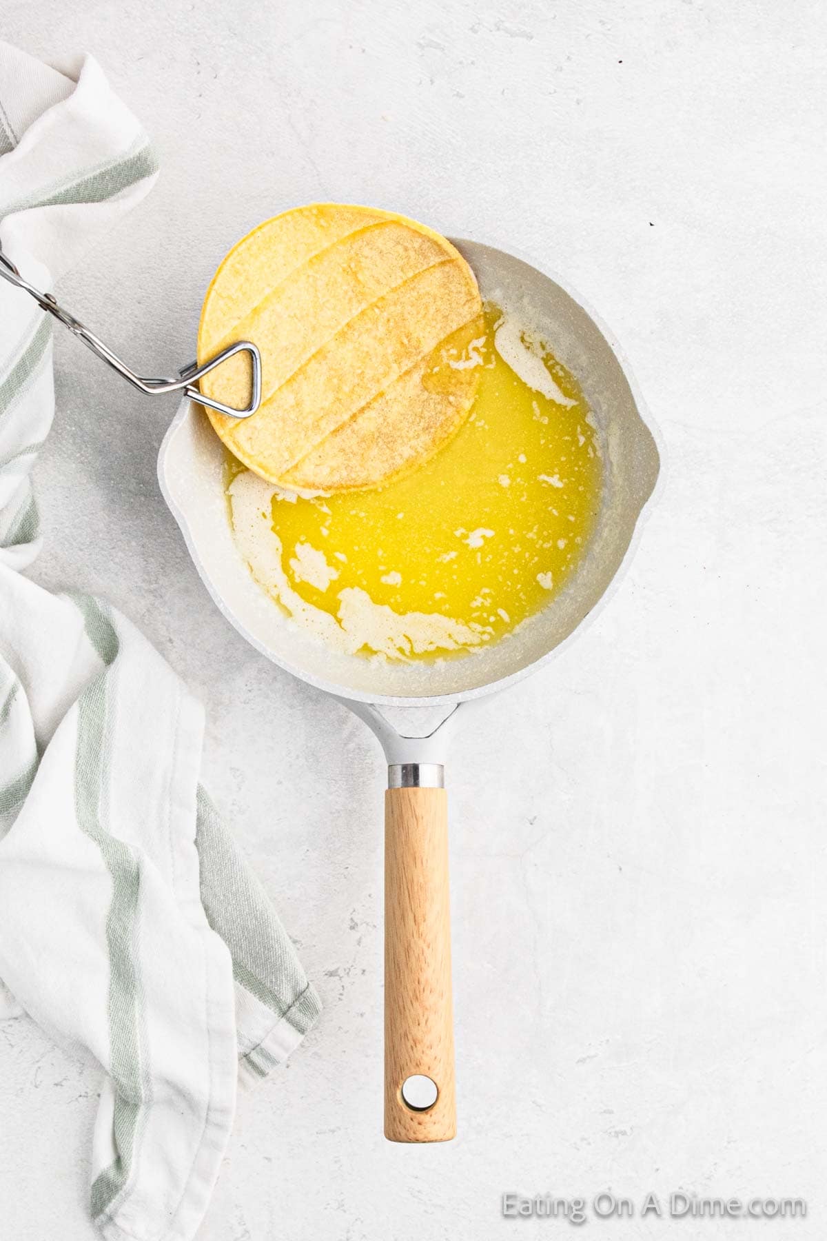 A yellow corn tortilla is being dipped into melted butter in a frying pan with a wooden handle, perfect for an easy fish tacos recipe. A white cloth with green stripes is placed beside the pan on a white surface.