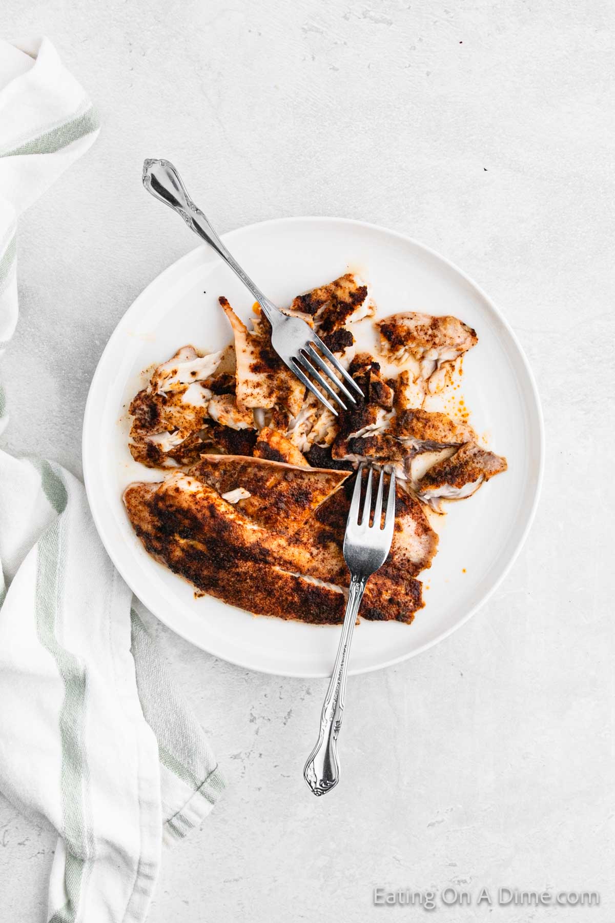 A white plate with blackened fish fillets, partially eaten, is placed on a light surface. Two forks rest on the plate. A white and green striped cloth napkin is partially visible on the left side of the image—an enticing visual for anyone searching for an easy fish tacos recipe.