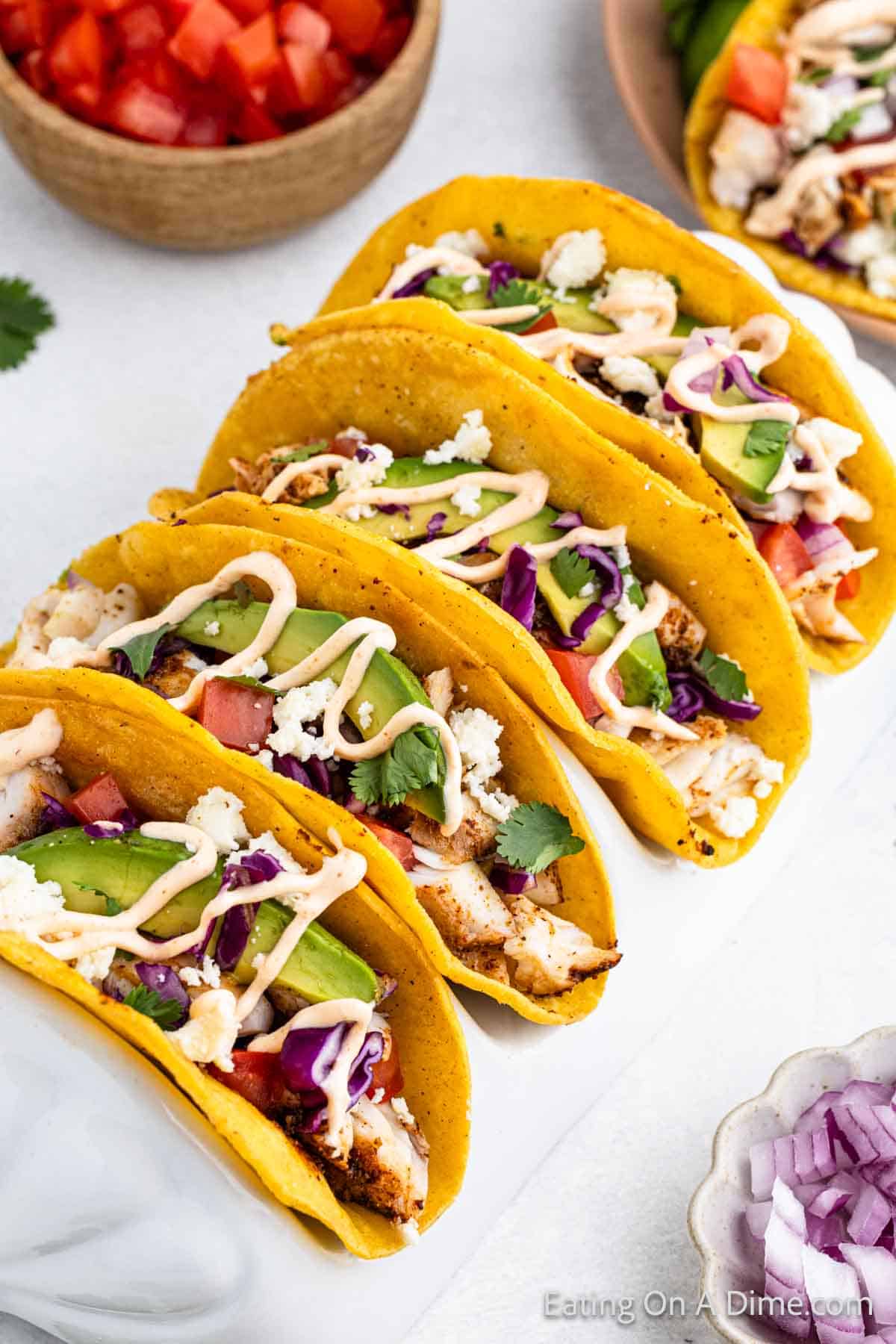 A plate of colorful fish tacos with ingredients including grilled fish, sliced avocado, diced tomatoes, shredded purple cabbage, cilantro, crumbled cheese, and a drizzle of creamy sauce in yellow corn tortillas. An easy fish tacos recipe, with a bowl of diced tomatoes seen in the background.