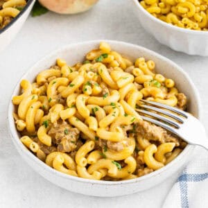 A white bowl filled with creamy macaroni and cheese, reminiscent of Homemade Cheeseburger Helper, garnished with chopped herbs. A fork is placed in the bowl, ready to scoop some of the pasta. A blue and white striped napkin is partially visible on the right side. Another bowl of plain macaroni is in the background.