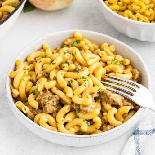 A white bowl filled with creamy macaroni and cheese, reminiscent of Homemade Cheeseburger Helper, garnished with chopped herbs. A fork is placed in the bowl, ready to scoop some of the pasta. A blue and white striped napkin is partially visible on the right side. Another bowl of plain macaroni is in the background.
