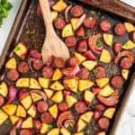 A baking sheet filled with Sausage and Potatoes, red onion pieces, all seasoned and mixed together. A wooden spoon stirs the mixture while fresh herbs are sprinkled on top for a flavorful finish.