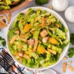 A bowl of Panera Bread Asian Sesame Chicken Salad Recipe features chopped lettuce, grilled chicken, mandarin orange slices, crispy wonton strips, and slivered almonds, arranged on a table with napkins and utensils nearby.