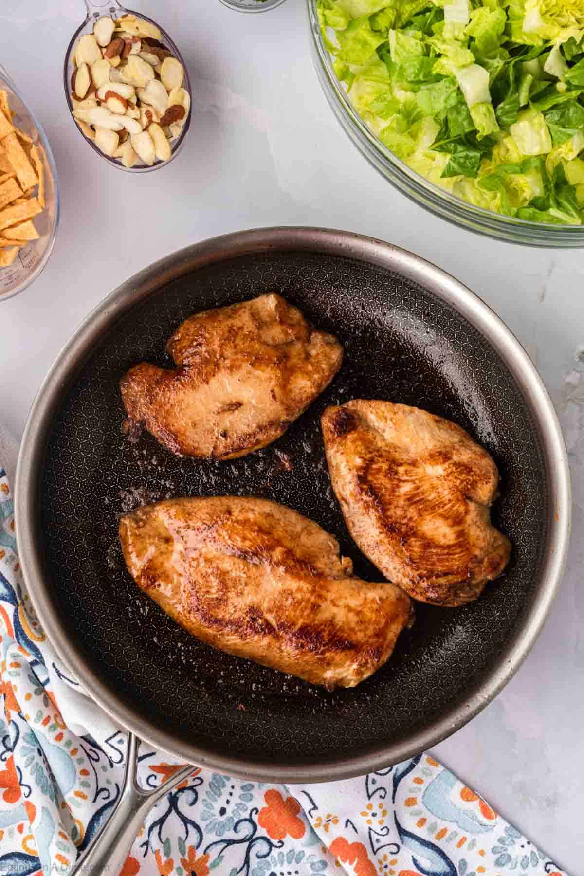 Cooking chicken in a skillet with a bowl of shredded romain, almonds