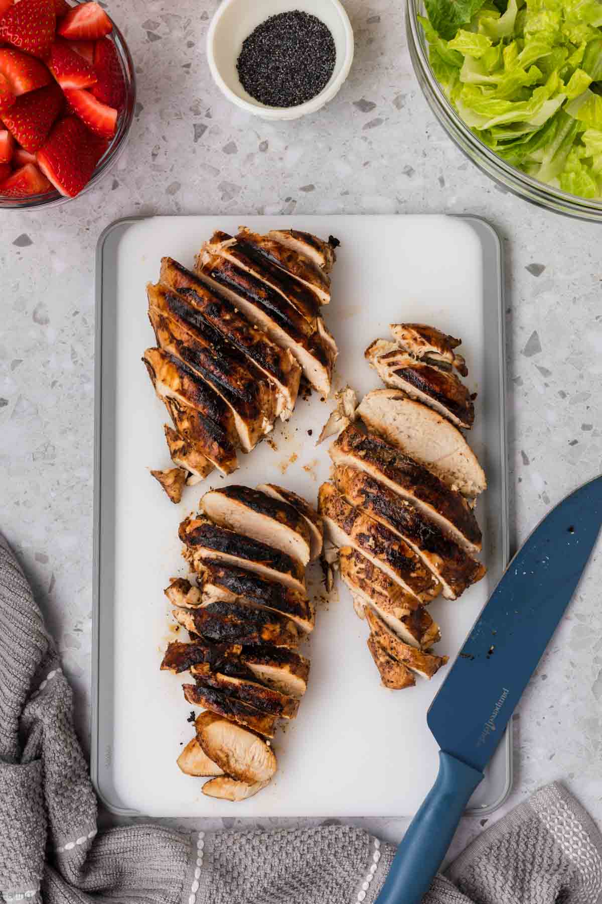 Sliced grilled chicken on a cutting board with a bowl of strawberries, poppyseed, and chopped lettuce