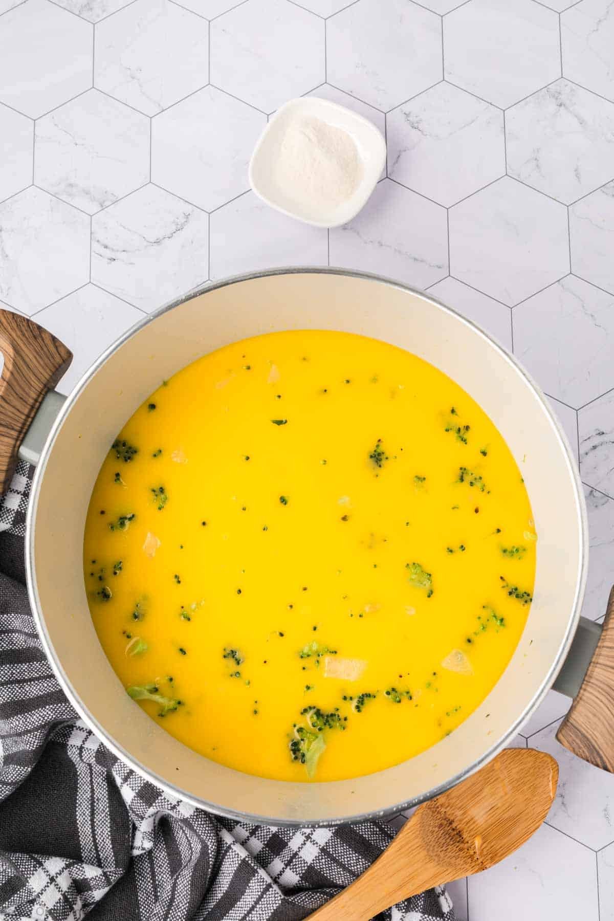 A large pot of Keto Broccoli Cheese Soup, filled with yellow soup, pieces of broccoli, and black pepper, sits on a checkered cloth. A wooden spoon rests nearby and a small dish of white powder is on the white hexagonal countertop.