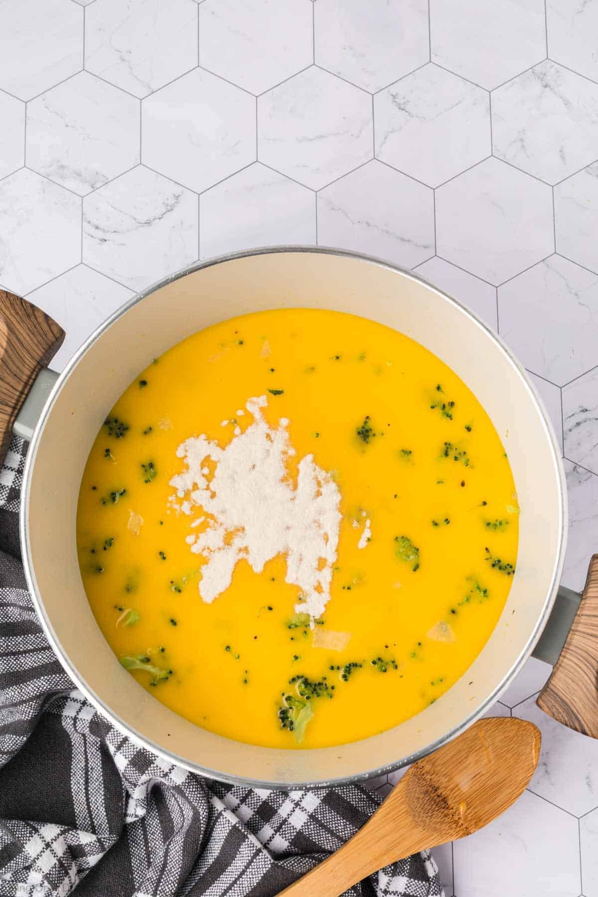 A pot of Keto Broccoli Cheese Soup with broccoli pieces and a pile of white powder on top sits on a marble-patterned surface, accompanied by a striped kitchen towel and wooden spoon.