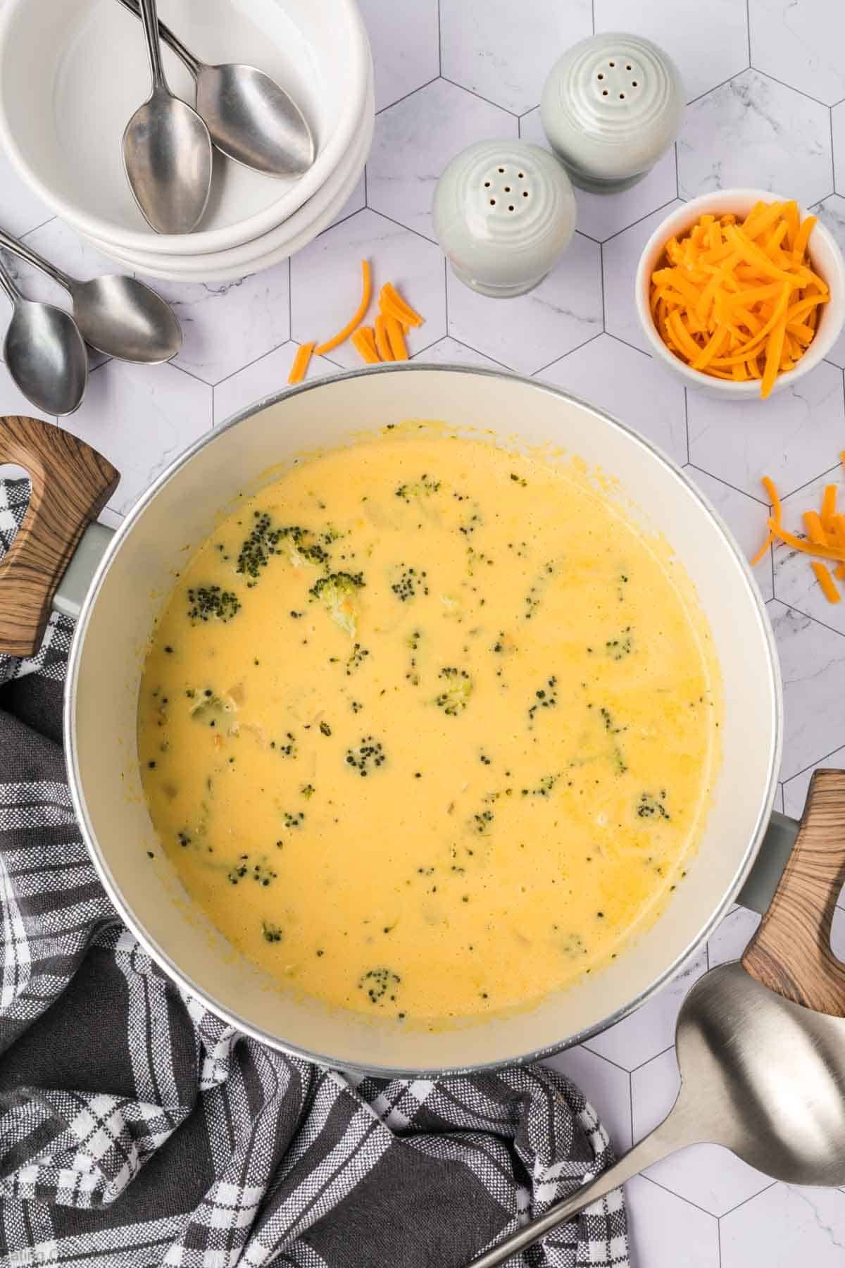 A pot of creamy Keto Broccoli Cheese Soup sits on a checkered towel, surrounded by a ladle, shredded cheese, spoons, stacked bowls, and salt and pepper shakers on a light hexagonal-tiled surface.