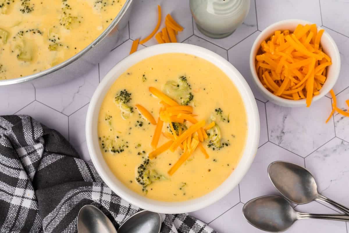 A bowl of creamy Keto Broccoli Cheese Soup topped with shredded cheddar cheese sits next to a bowl of extra cheese, a checkered towel, two spoons, and a pot of soup on a white hexagon tile surface.