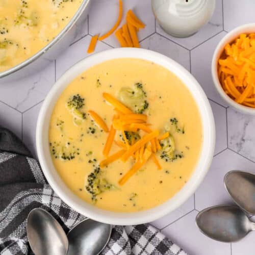 A bowl of creamy Keto Broccoli Cheese Soup topped with shredded cheddar cheese sits on a white hexagonal-tiled surface, next to a bowl of more shredded cheese, a checkered napkin, and three metal spoons.