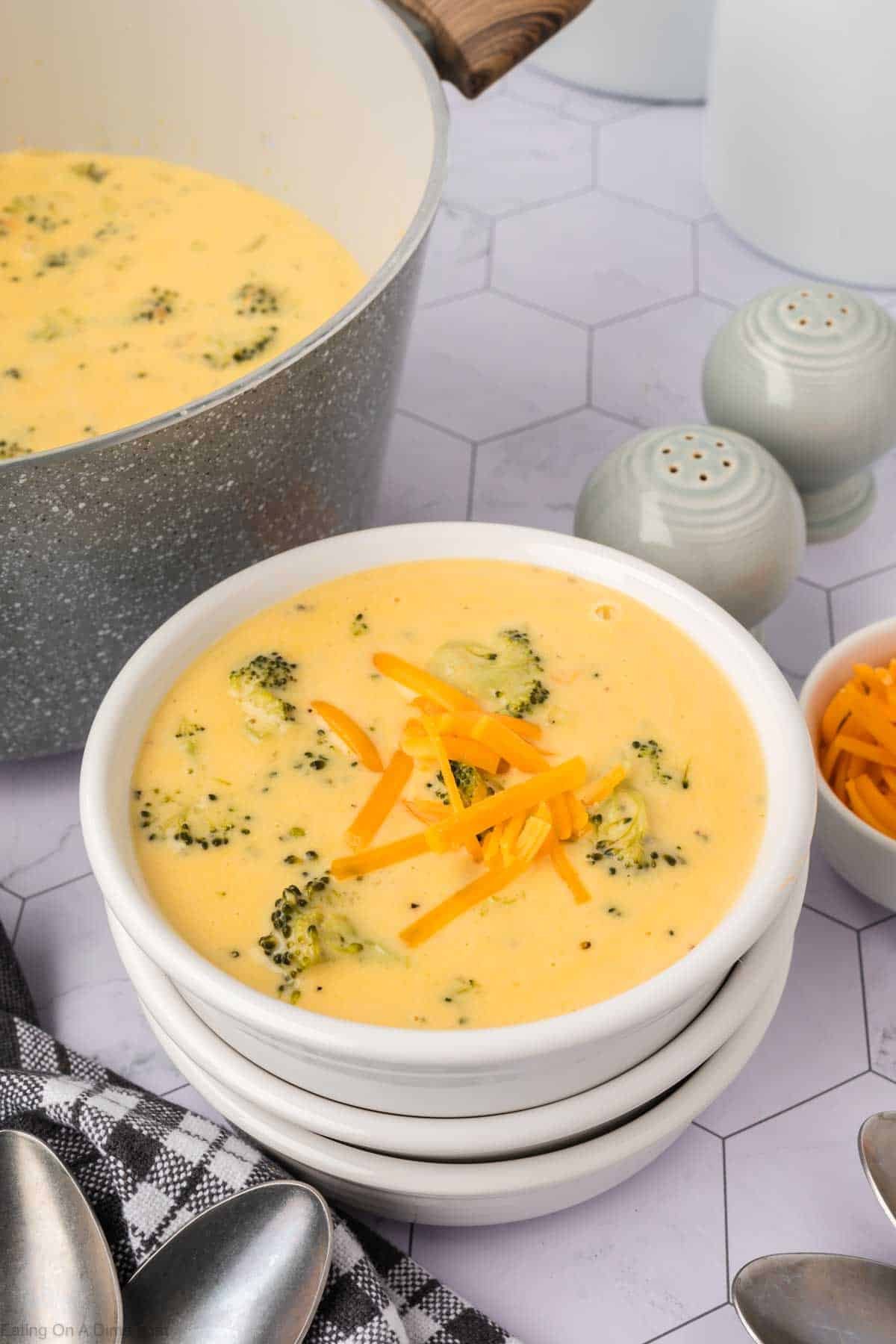 A bowl of creamy Keto Broccoli Cheese Soup topped with shredded cheddar sits on stacked white bowls. Nearby are a pot of soup, extra cheese, salt and pepper shakers, and a checkered napkin with spoons.