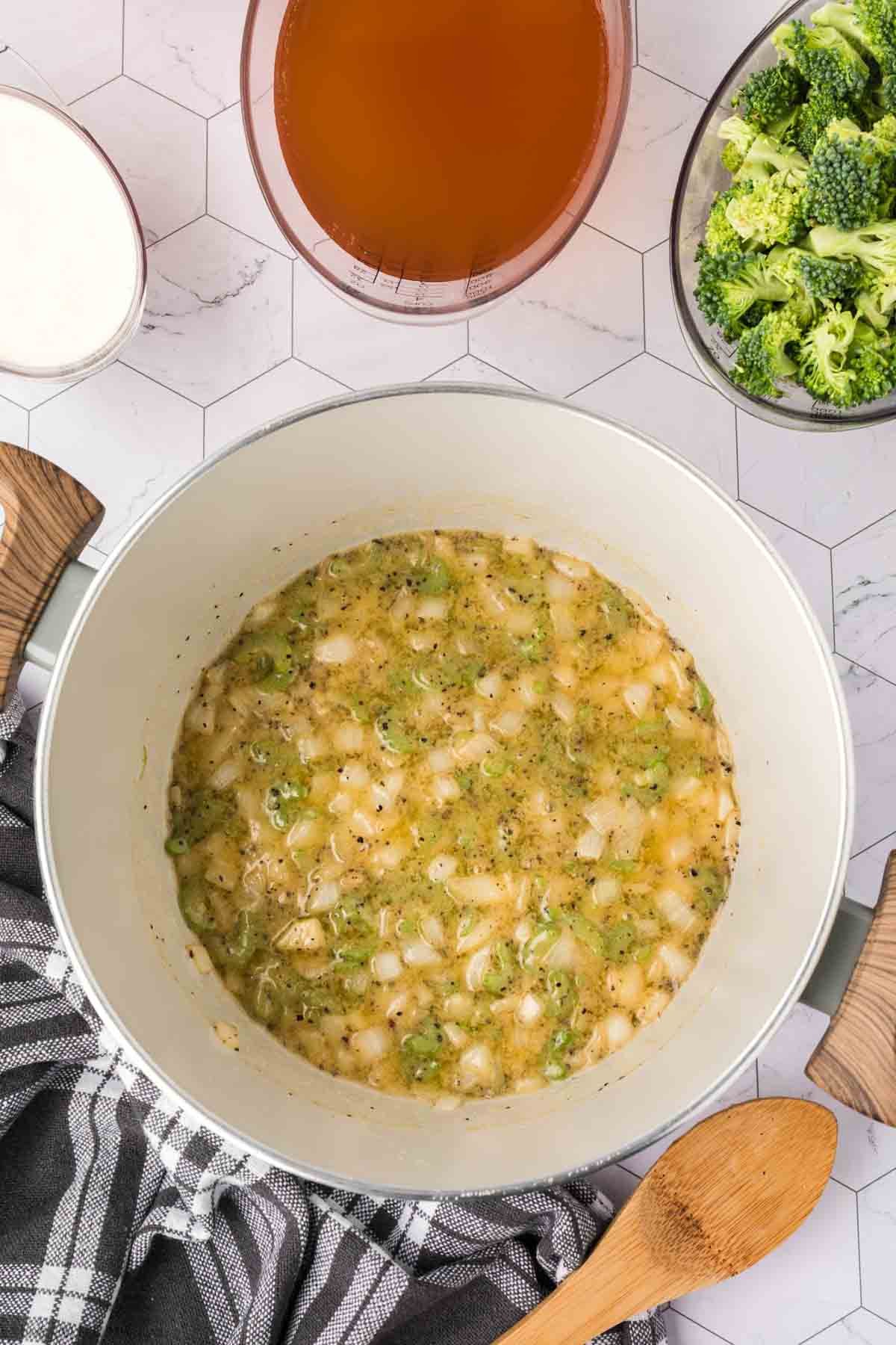 A pot of sautéed onions and seasonings sits on a counter, surrounded by bowls of broccoli florets, broth, and cream—perfect ingredients for a comforting Keto Broccoli Cheese Soup. A wooden spoon rests nearby on a folded kitchen towel.