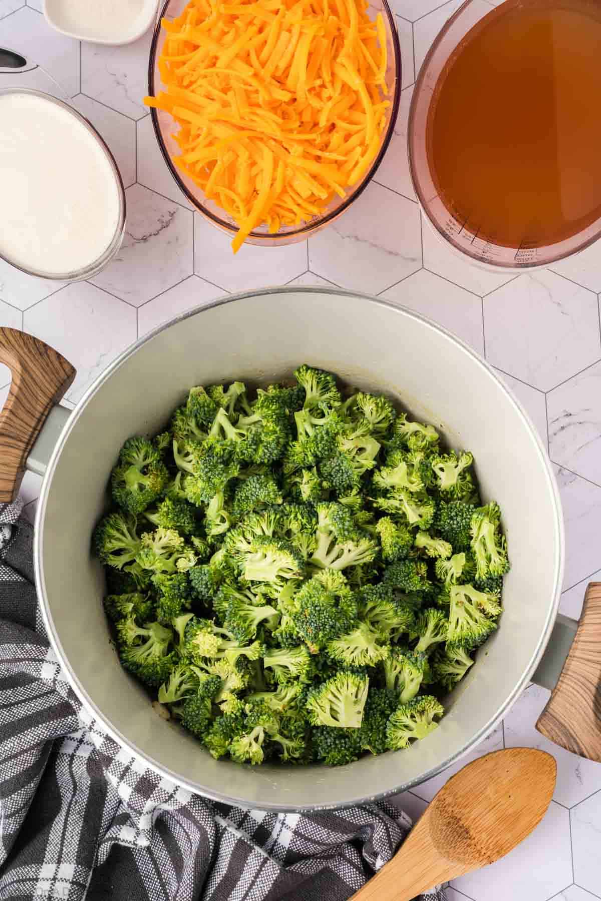 A pot filled with fresh broccoli florets sits on a patterned countertop, surrounded by bowls of shredded cheddar cheese, broth, and cream—everything you need to make delicious Keto Broccoli Cheese Soup—alongside a wooden spoon and checkered towel.
