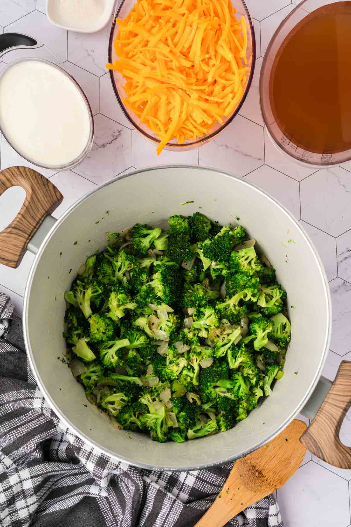 A pot filled with cooked broccoli and onions sits on a counter next to bowls of shredded cheddar cheese, broth, and cream—perfect ingredients for a hearty Keto Broccoli Cheese Soup. A wooden spoon and black-and-white kitchen towel are nearby.