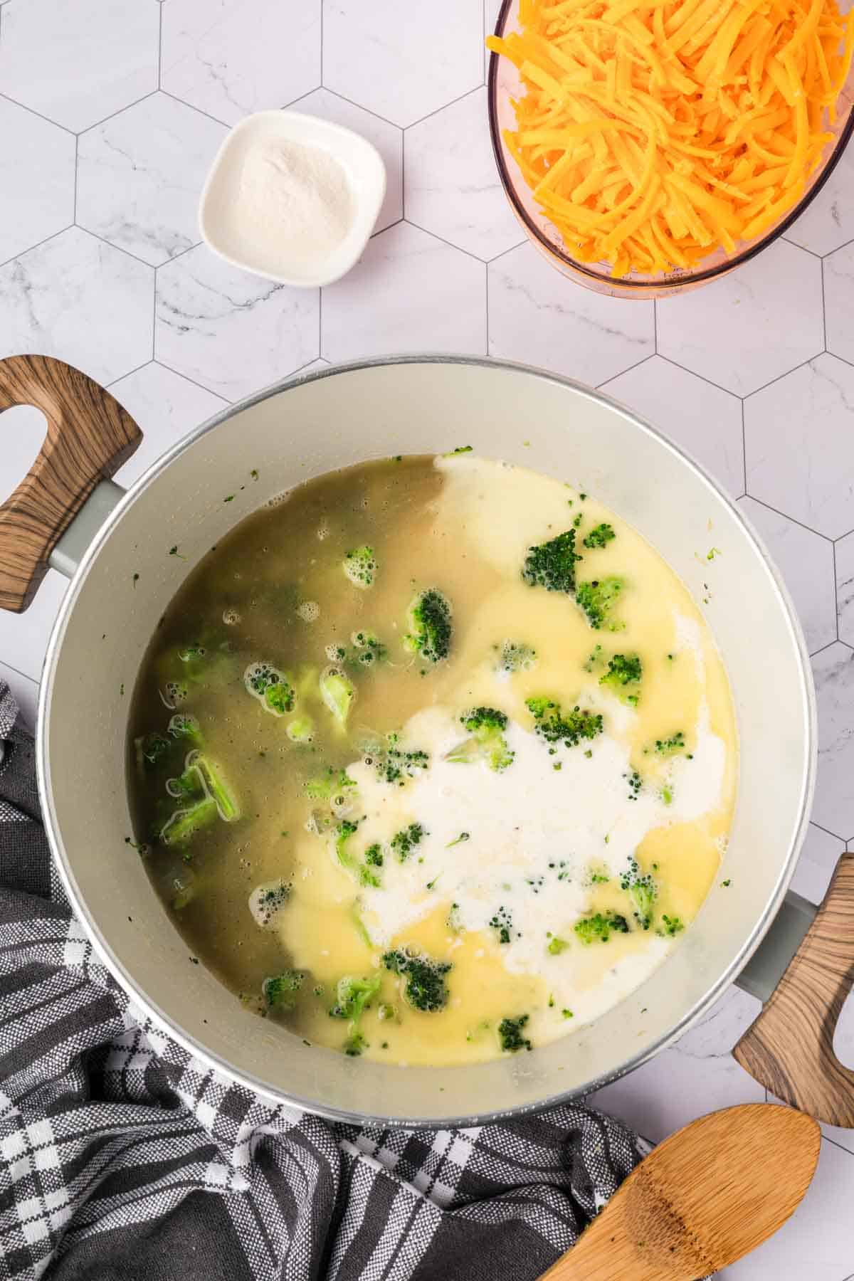 A pot of Keto Broccoli Cheese Soup, rich with broccoli, broth, and cream, sits on a marble surface next to a bowl of shredded cheddar, a dish of flour, a wooden spoon, and a black-and-white kitchen towel.