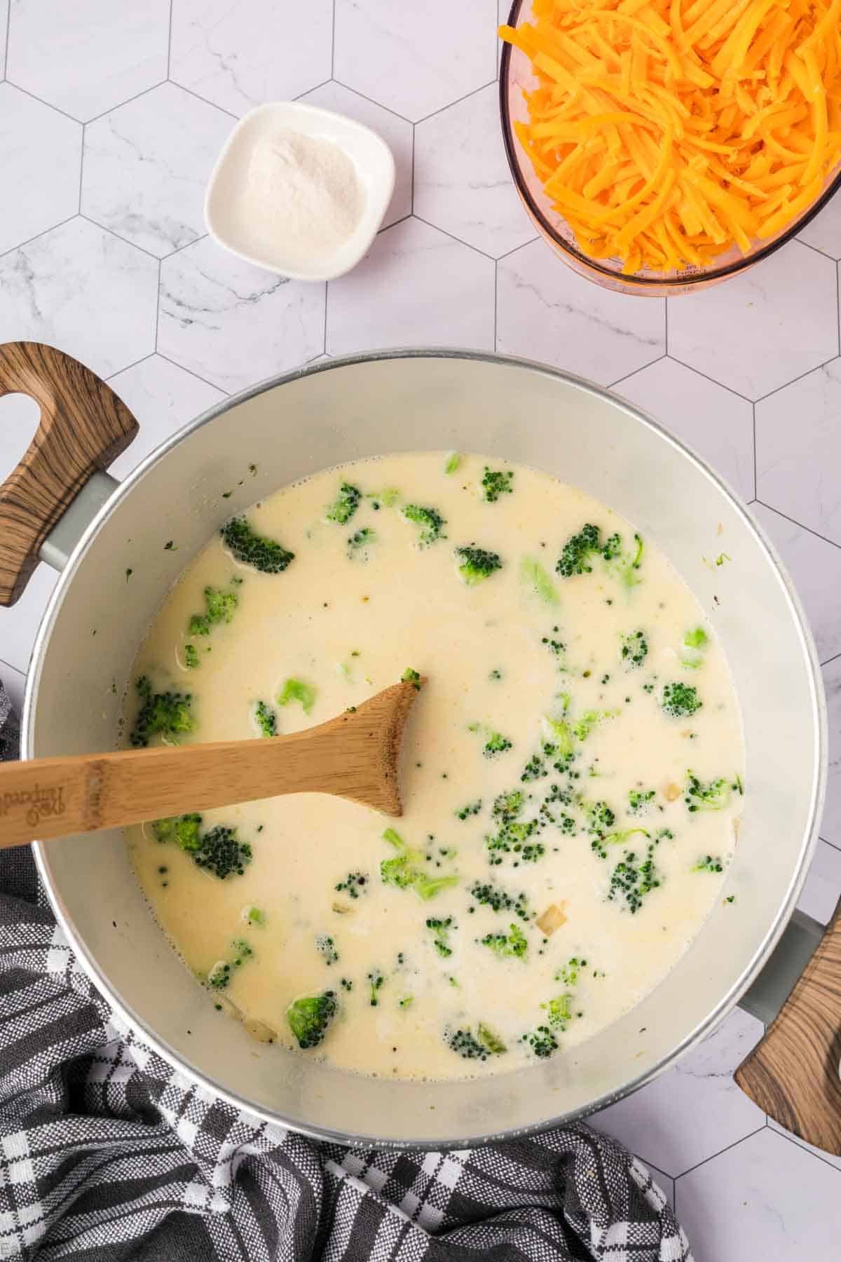 A pot of creamy Keto Broccoli Cheese Soup with broccoli florets is stirred with a wooden spoon, set beside shredded cheddar cheese and a small dish of white powder on a hexagon-tiled countertop.