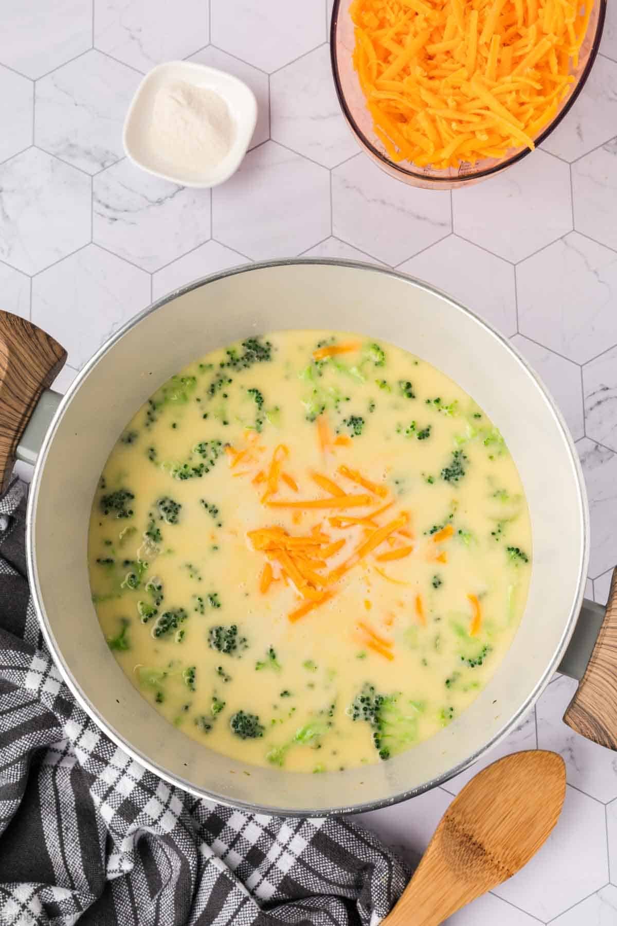 A pot of creamy Keto Broccoli Cheese Soup with cheese sprinkled on top, sitting on a white hexagon-tiled surface next to a bowl of shredded cheddar, a small dish of grated cheese, and a wooden spoon on a striped towel.