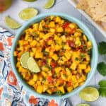 A bowl of vibrant Mango Salsa with diced mango, red onion, red pepper, cilantro, and lime is garnished with lime wedges. Tortilla chips, lime slices, and a patterned cloth complete this fresh and zesty spread.