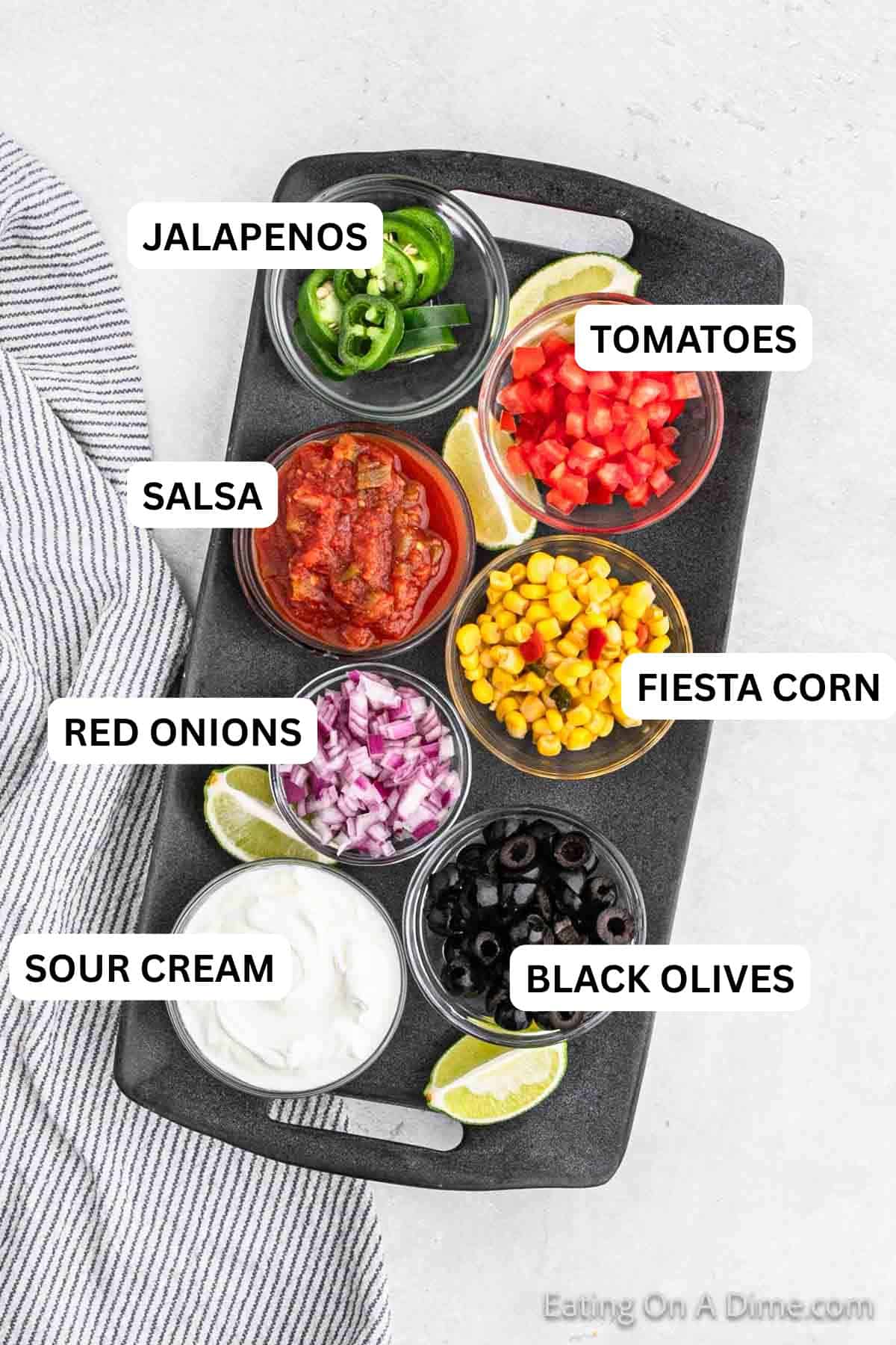 A black serving tray showcases small bowls of sliced jalapeños, salsa, diced tomatoes, corn, red onions, black olives, and sour cream. Lime wedges sit beside the bowls. A striped napkin is partially visible to the left of the tray.
