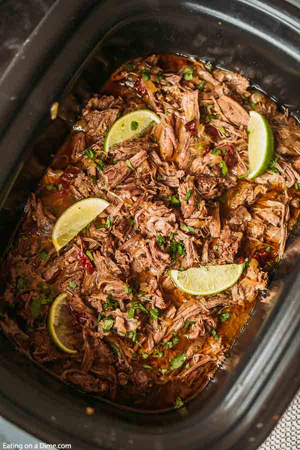 Close up image of Barbacoa Beef in the crock pot with cilantro on top with limes. 