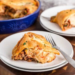 A slice of cheesy, baked lasagna on a white plate with a fork, with another plate of lasagna and an Italian Crescent Casserole in a blue dish visible in the background.