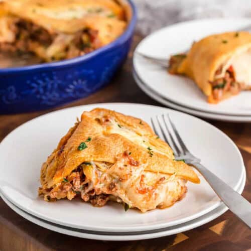 A slice of cheesy, baked lasagna on a white plate with a fork, with another plate of lasagna and an Italian Crescent Casserole in a blue dish visible in the background.