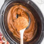 A white spoon holds a scoop of thick, orange-brown pumpkin butter over a slow cooker, showcasing this delicious Crock Pot Pumpkin Butter Recipe. A red-and-white cloth is partially visible beside the slow cooker.