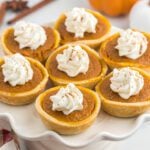 Mini pumpkin pies topped with swirls of whipped cream are arranged on a white scalloped cake stand. Featuring golden crusts and a sprinkle of spice, this mini pumpkin pies recipe creates a festive dessert display.