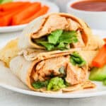 A close-up of a Buffalo Chicken Wraps Recipe, filled with lettuce, cut in half and stacked on a white plate, served with carrot sticks, celery, chips, and a bowl of dipping sauce in the background.