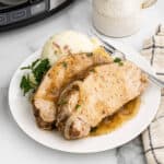 Two seasoned pork chops topped with onions and gravy, reminiscent of a comforting crock pot pork loin recipe, served with mashed potatoes and parsley. A fork rests beside a striped napkin and mug on the white plate.