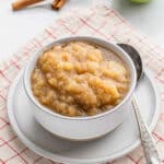 A white bowl of chunky crockpot applesauce sits on a white plate with a silver spoon, atop a red and white checkered cloth. Two cinnamon sticks are in the background.