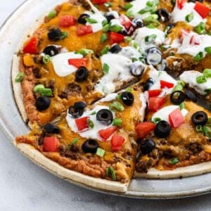 A pizza topped with melted cheese, ground meat, sliced black olives, chopped tomatoes, green onions, and dollops of sour cream on a metal tray—this taco pizza recipe even has one slice slightly pulled out from the rest.