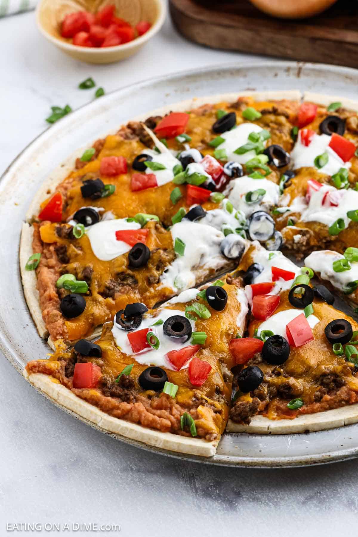 A delicious taco pizza recipe featuring a baked Mexican pizza topped with ground beef, melted cheese, black olives, tomatoes, green onions, and dollops of sour cream, all served on a round metal tray.