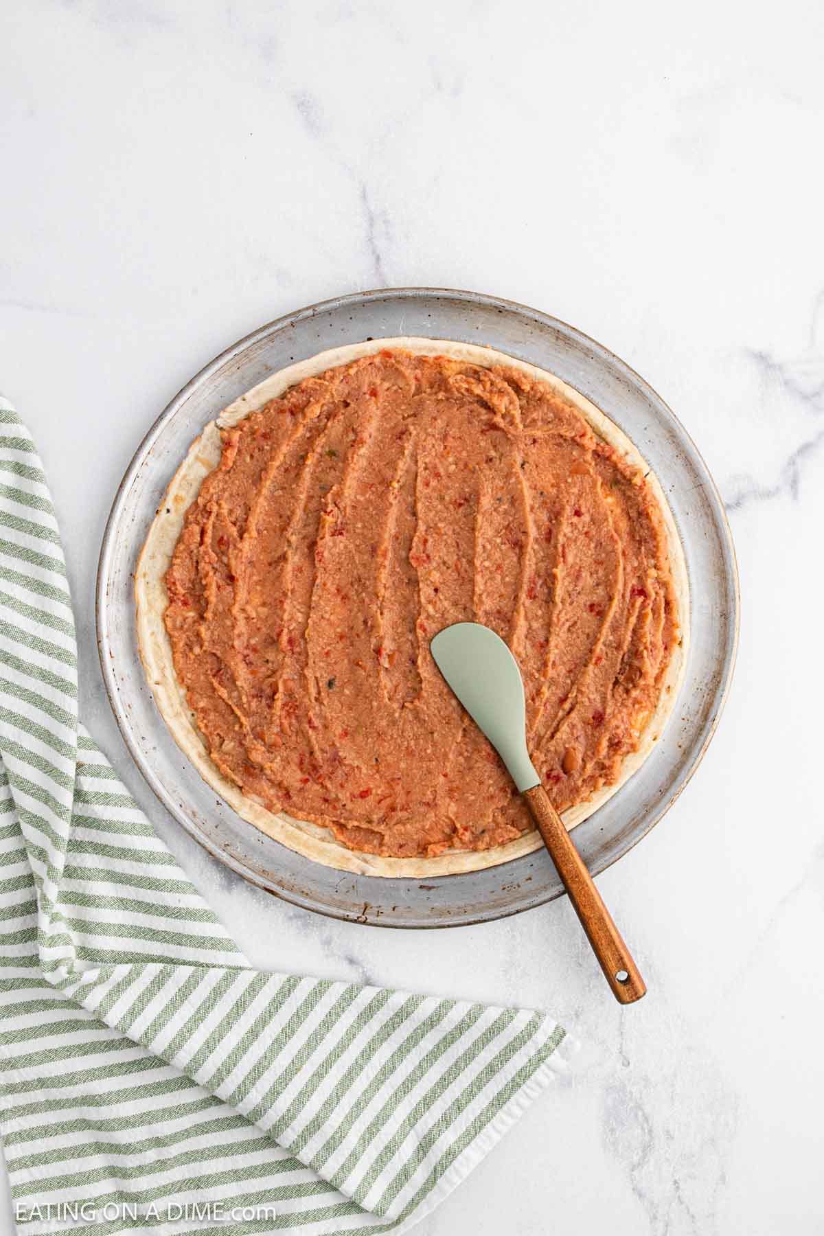 A pizza crust spread with a layer of refried beans, as part of a taco pizza recipe, shown on a round plate with a green-striped kitchen towel beside it and a green spatula resting on the beans.