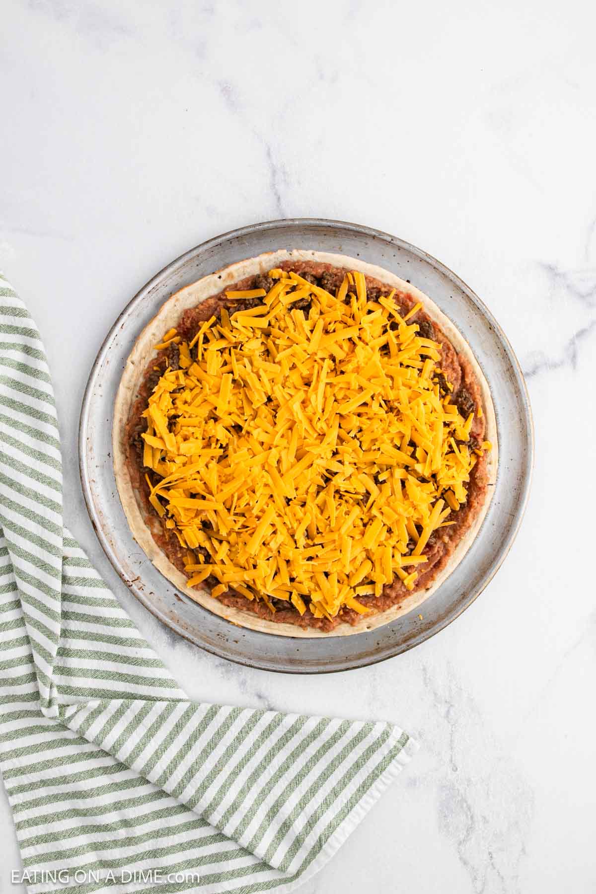 A round pizza crust topped with shredded cheddar cheese and a layer of beans, this taco pizza recipe is served on a plate with a green and white striped cloth nearby on a marble surface.